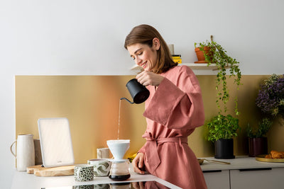 Woman in a pink robe pouring coffee in a kitchen with plants and a yellow wall.