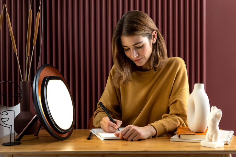 Woman sitting at a desk with a light therapy lamp, notebook, and pen.